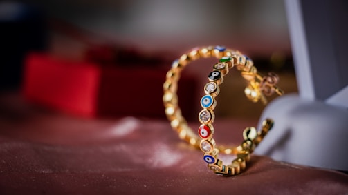 A close-up of a pair of gold earrings adorned with colorful enamel designs and small gemstones. The earrings are resting on a soft pink fabric surface with a blurred red box in the background. The focus is on the intricate detailing of the jewelry, highlighting the vibrant decorative elements.