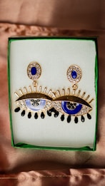 A pair of earrings in the shape of stylized eyes with gold detailing and blue accents are displayed in a green-bordered box. The eyes have an intricate design with eyelashes and feature detailed embellishments with small stones surrounding them.