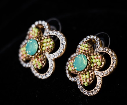 Two ornate earrings with a floral design featuring a central turquoise stone surrounded by small multicolored beads. Each petal-like section is bordered by a series of clear gemstones, creating a sparkling effect against a dark background.