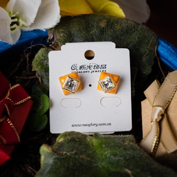 A pair of orange earrings with clear gemstones presented on a white card from Neoglory Jewelry. The card rests on a dark green surface surrounded by fabric flowers and a gold-accented red gift box ribbon, creating a rich and elegant display.