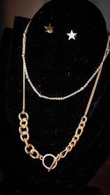 A display of a gold necklace with a layered chain and star-shaped earrings is elegantly arranged on a black velvet stand. The necklace features a combination of thin and chunky links, creating a stylish contrast.