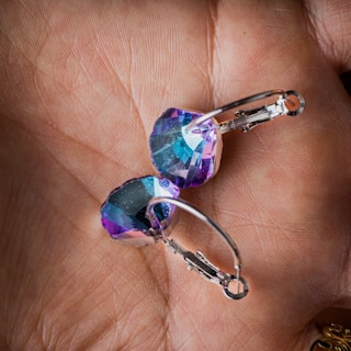 A pair of earrings with faceted, colorful gemstones held in a hand. The gemstones have a combination of blue, purple, and teal colors, and the earrings have metal clasps.