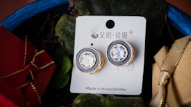 A pair of elegant earrings with large, clear stones set in gold-colored metal are displayed on a white card. The card has decorative patterns and text. The earrings are surrounded by greenery and colorful gift boxes, adding a festive touch to the scene.