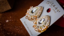 A pair of ornate earrings with a teardrop shape, adorned with sparkling crystals and beads. The earrings rest on a card with elegant branding and are positioned against a textured brown surface, creating a contrast between the shiny jewelry and the rich background.