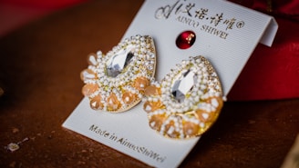 A pair of decorative earrings placed on a display card. They feature intricate designs with shiny gems and beads, including large clear central stones and smaller orange and white beads. The card displays some text in both English and another language, along with the brand name 'Ainuo Shiwei'.