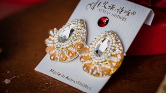 A pair of decorative earrings placed on a display card. They feature intricate designs with shiny gems and beads, including large clear central stones and smaller orange and white beads. The card displays some text in both English and another language, along with the brand name 'Ainuo Shiwei'.