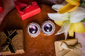 A pair of elegant earrings with large purple gemstones surrounded by intricate gold and blue settings lies on a textured surface. Nearby, there are several small gift boxes wrapped in red and brown paper with decorative ribbons. Yellow and white flowers are placed on the right side, adding a touch of natural beauty to the scene.