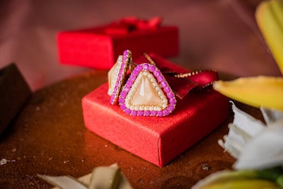 A pair of triangular earrings with pearl and pink stone accents is elegantly placed on a vibrant red gift box. The textured surface of the box adds a touch of luxury to the overall presentation. The background includes soft-focus elements and hints of fabric, complementing the setup.