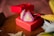 A pair of triangular earrings with pearl and pink stone accents is elegantly placed on a vibrant red gift box. The textured surface of the box adds a touch of luxury to the overall presentation. The background includes soft-focus elements and hints of fabric, complementing the setup.