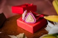 A pair of triangular earrings with pearl and pink stone accents is elegantly placed on a vibrant red gift box. The textured surface of the box adds a touch of luxury to the overall presentation. The background includes soft-focus elements and hints of fabric, complementing the setup.