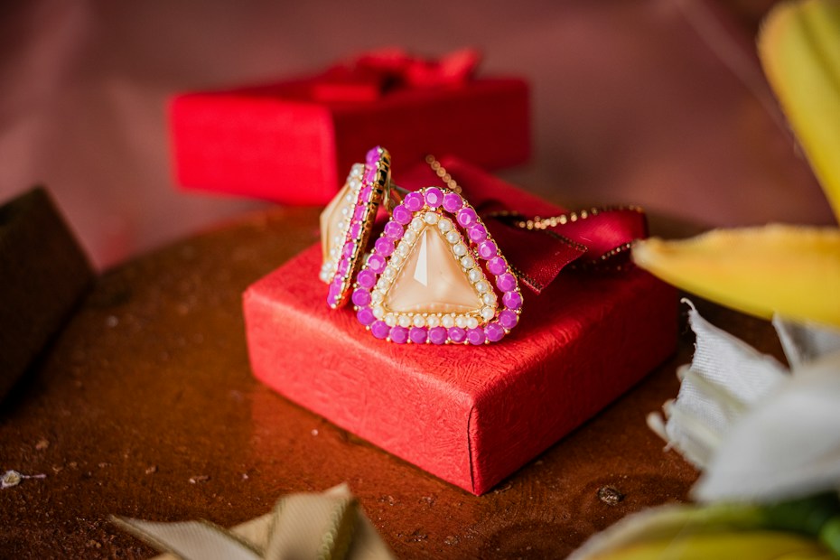 A pair of triangular earrings with pearl and pink stone accents is elegantly placed on a vibrant red gift box. The textured surface of the box adds a touch of luxury to the overall presentation. The background includes soft-focus elements and hints of fabric, complementing the setup.