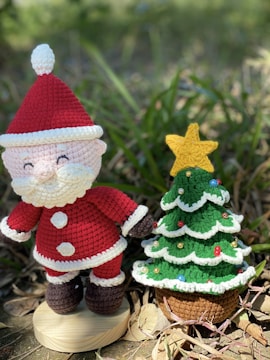 A crochet Santa Claus figure stands next to a crocheted Christmas tree decorated with colorful beads. The scene is set outdoors on a natural surface with green foliage in the background.