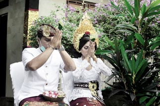 A man and a woman dressed in traditional attire are sitting with their hands pressed together in a praying gesture. They are surrounded by lush greenery and flowering plants. The woman wears an elaborate headdress and the man wears a traditional hat.