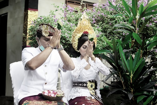 A man and a woman dressed in traditional attire are sitting with their hands pressed together in a praying gesture. They are surrounded by lush greenery and flowering plants. The woman wears an elaborate headdress and the man wears a traditional hat.