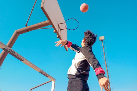 A person attempts a basketball shot outdoors. The hoop is visible with the basketball in mid-air. The sky is clear and blue, and the person is wearing a black and white sweatshirt.