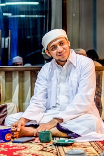 A man dressed in traditional white attire with a headscarf is sitting on a decorative carpet indoors. He appears relaxed, with his hands resting on his legs. There is a cup and a small container on the floor in front of him. The setting includes a large window reflecting the interior lights.
