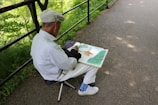 An elderly person, wearing a flat cap and a light-colored shirt, is seated on a small folding stool by a paved pathway. They are absorbed in sketching or painting in a large sketchbook. The surroundings are lush with green foliage, and the path is bordered by a black metal railing.