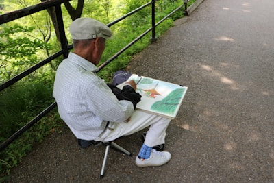 An elderly person, wearing a flat cap and a light-colored shirt, is seated on a small folding stool by a paved pathway. They are absorbed in sketching or painting in a large sketchbook. The surroundings are lush with green foliage, and the path is bordered by a black metal railing.