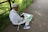 An elderly person, wearing a flat cap and a light-colored shirt, is seated on a small folding stool by a paved pathway. They are absorbed in sketching or painting in a large sketchbook. The surroundings are lush with green foliage, and the path is bordered by a black metal railing.