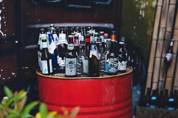 A collection of empty beer bottles is placed on top of a red metal barrel. The barrels and the bottles show various brands and labels. In the background, there is a green wall with bamboo stalks leaning against it and additional bottles scattered on the floor.