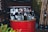 A collection of empty beer bottles is placed on top of a red metal barrel. The barrels and the bottles show various brands and labels. In the background, there is a green wall with bamboo stalks leaning against it and additional bottles scattered on the floor.