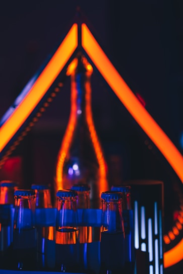 A collection of glass bottles with glowing blue and orange accents creates a visually striking display. The geometric shape formed by vibrant orange neon lights in the background adds an artistic and modern aesthetic.