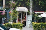 A quaint vintage store entrance is nestled between lush greenery and vines. The shop has a charming sign above the door and displays various items inside. A bicycle is partially visible outside, and there are small decorative plants and a white patio umbrella nearby.