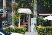 A quaint vintage store entrance is nestled between lush greenery and vines. The shop has a charming sign above the door and displays various items inside. A bicycle is partially visible outside, and there are small decorative plants and a white patio umbrella nearby.
