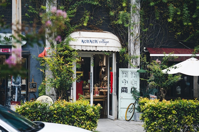A quaint vintage store entrance is nestled between lush greenery and vines. The shop has a charming sign above the door and displays various items inside. A bicycle is partially visible outside, and there are small decorative plants and a white patio umbrella nearby.