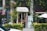 A quaint vintage store entrance is nestled between lush greenery and vines. The shop has a charming sign above the door and displays various items inside. A bicycle is partially visible outside, and there are small decorative plants and a white patio umbrella nearby.