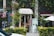 A quaint vintage store entrance is nestled between lush greenery and vines. The shop has a charming sign above the door and displays various items inside. A bicycle is partially visible outside, and there are small decorative plants and a white patio umbrella nearby.