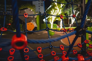 A nighttime playground scene with a climbing net featuring blue ropes and red connectors in the foreground. In the background, there are illuminated trees with white lights and colorful playground equipment, including a geometric climbing structure.