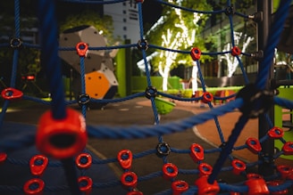 A nighttime playground scene with a climbing net featuring blue ropes and red connectors in the foreground. In the background, there are illuminated trees with white lights and colorful playground equipment, including a geometric climbing structure.