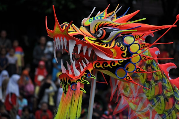 A vibrant and intricately designed dragon puppet with vivid colors and sharp features is being held up in a lively outdoor setting. The background is filled with a crowd of spectators, adding a sense of festivity.