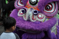 A young child stands in front of a large, vibrant, purple lion dance costume, adorned with intricate decorations in red, white, and black. The lion costume has a fluffy texture and large, expressive eyes, capturing the child's attention.
