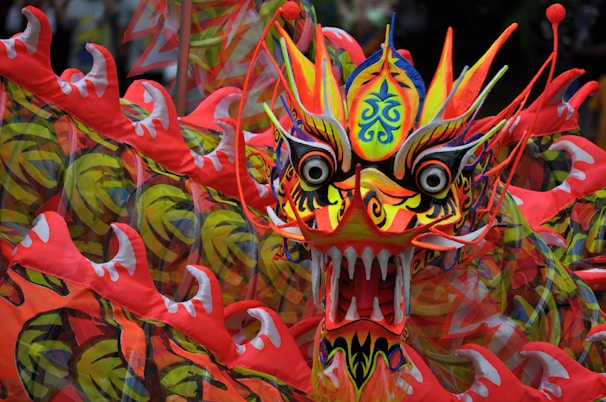 A vibrant and intricately designed dragon head with bold colors and detailed patterns. The dragon features sharp teeth, large eyes, and various swirling designs in its skin, with red, yellow, and orange hues dominating its appearance.