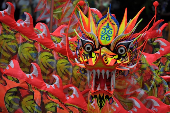 A vibrant and intricately designed dragon head with bold colors and detailed patterns. The dragon features sharp teeth, large eyes, and various swirling designs in its skin, with red, yellow, and orange hues dominating its appearance.