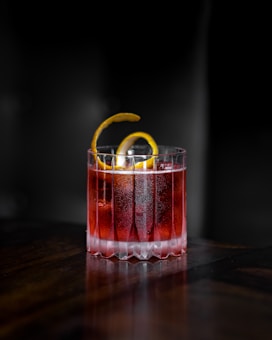 A glass filled with a reddish cocktail sits on a dark wooden surface. The drink is garnished with a twisted lemon peel, adding a touch of elegance. The lighting creates a striking contrast, highlighting the textured glass and the vibrant hue of the beverage.