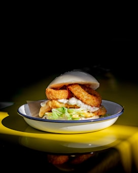 A sandwich with a soft bun filled with crispy fried onions, lettuce, and sauce is placed on a white plate with a blue rim. The background is dark, highlighting the sandwich, and the image has a bright reflection of yellow on the surface.