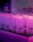 Frosted glass mugs with black geometric symbols are arranged on a black rubber mat atop a white tiled surface. The scene is lit with a pink and purple neon glow, giving a vibrant, atmospheric look.