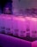 Frosted glass mugs with black geometric symbols are arranged on a black rubber mat atop a white tiled surface. The scene is lit with a pink and purple neon glow, giving a vibrant, atmospheric look.