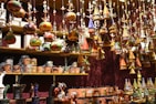 A variety of colorful, handcrafted ornaments and ceramics are displayed on shelves and hanging from above. The ornaments include small houses with conical roofs and various whimsical shapes with intricate patterns. The shelves are filled with mugs and other pottery pieces, some wrapped in protective material.
