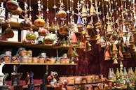 A variety of colorful, handcrafted ornaments and ceramics are displayed on shelves and hanging from above. The ornaments include small houses with conical roofs and various whimsical shapes with intricate patterns. The shelves are filled with mugs and other pottery pieces, some wrapped in protective material.
