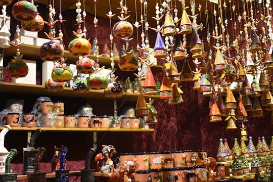 A variety of colorful, handcrafted ornaments and ceramics are displayed on shelves and hanging from above. The ornaments include small houses with conical roofs and various whimsical shapes with intricate patterns. The shelves are filled with mugs and other pottery pieces, some wrapped in protective material.