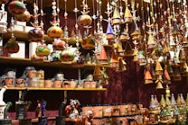 A variety of colorful, handcrafted ornaments and ceramics are displayed on shelves and hanging from above. The ornaments include small houses with conical roofs and various whimsical shapes with intricate patterns. The shelves are filled with mugs and other pottery pieces, some wrapped in protective material.