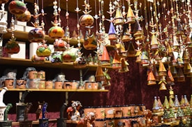 A variety of colorful, handcrafted ornaments and ceramics are displayed on shelves and hanging from above. The ornaments include small houses with conical roofs and various whimsical shapes with intricate patterns. The shelves are filled with mugs and other pottery pieces, some wrapped in protective material.