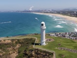 A white lighthouse stands prominently on a grassy cliff overlooking the ocean. The coastline stretches into the distance with clear turquoise waters meeting a sandy beach. Numerous small waves roll toward the shore, where many people are swimming and sunbathing. In the background, a cityscape with industrial buildings and smokestacks is visible. A parking area filled with cars is adjacent to the lighthouse.