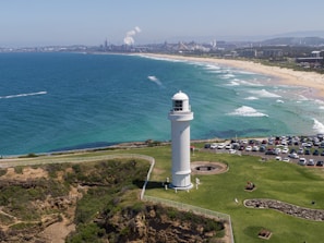 A white lighthouse stands prominently on a grassy cliff overlooking the ocean. The coastline stretches into the distance with clear turquoise waters meeting a sandy beach. Numerous small waves roll toward the shore, where many people are swimming and sunbathing. In the background, a cityscape with industrial buildings and smokestacks is visible. A parking area filled with cars is adjacent to the lighthouse.