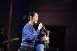 A person is singing passionately into a microphone on a dimly lit stage. The individual is wearing a blue sweater and has closed eyes, suggesting they are deeply focused or moved by the music. In the background, another person can be seen holding a microphone with blurred surroundings, emphasizing the main subject.