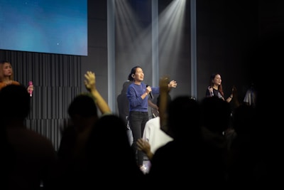 A singer in a blue shirt is performing on stage under bright spotlights, with other performers in the background. The audience is visible in the foreground with raised hands, creating an atmosphere of engagement and excitement. The setting is likely a concert or live performance.